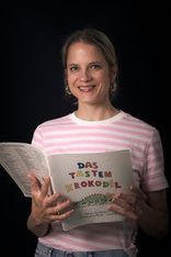 Eine Frau in einem rosa-weiß gestreiften T-Shirt hält ein offenes Buch mit dem Titel 'Das Tasten Krokodil' und lächelt in die Kamera.
