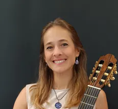 Eine Frau mit langen, gewellten braunen Haaren lächelt und posiert für ein Foto, während sie eine Gitarre hält.