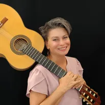 Eine Frau mit kurzen grauen Haaren lächelt und hält eine Gitarre.