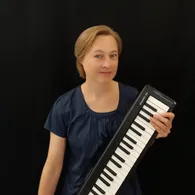 Eine Frau mit kurzen blonden Haaren lächelt und hält ein schwarzes und weißes Keyboard. Sie trägt ein blaues Oberteil.