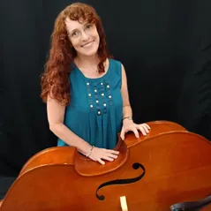 Eine Frau mit lockigem rotem Haar steht neben einem großen Cello und posiert für ein Foto.