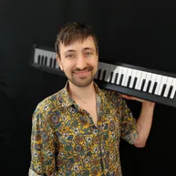 Ein Mann lächelt, während er eine Tastatur vor sich hält. Er trägt ein gelbes Blumenhemd und eine Halskette.