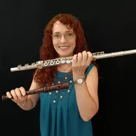 Eine Frau mit lockigem rotem Haar hält eine silberne Flöte und ein hölzerne Blockflöte in ihren Händen. Sie trägt ein blaues Oberteil und eine Uhr.