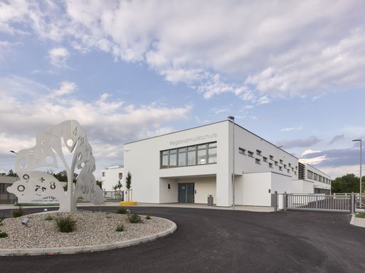 Ein modernes weißes Gebäude mit Glasfenstern und einem Schild, das 'Regionalmusikschule' besagt, befindet sich vor einem Kieskreisverkehr. Das Gebäude ist von einem Zaun und einem Baum umgeben. Der Himmel ist blau mit Wolken.
