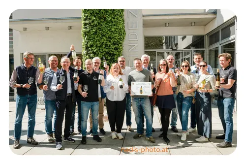 Eine Gruppe von Menschen posiert für ein Foto vor einem Gebäude mit dem Schriftzug 'Linden'. Sie halten alle Weingläser und Flaschen Wein.