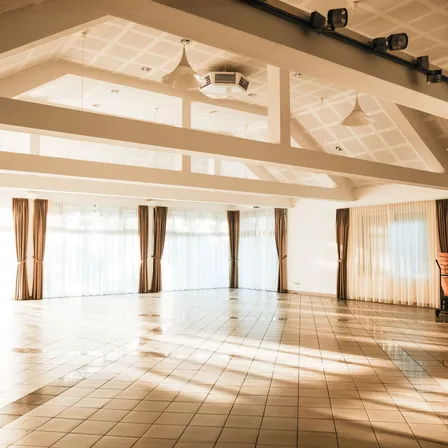Ein leerer Saal mit großen Fenstern und Vorhängen, einem weißen Fliesenboden und einer Klimaanlage an der Decke.