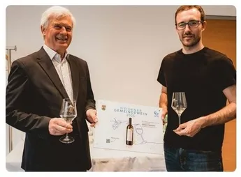 Zwei lächelnde Männer, einer hält ein Weinglas und der andere einen Weinflasche mit einem Schild, das 'Weidener Gemeindewein 2025' steht.