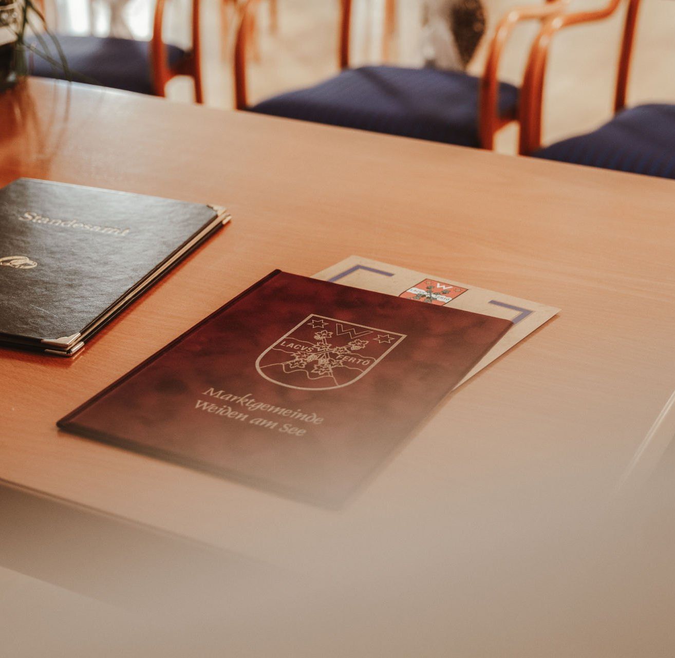 Zwei Bücher mit geprägten Logos auf einem Holztisch, mit leeren Stühlen im Hintergrund.