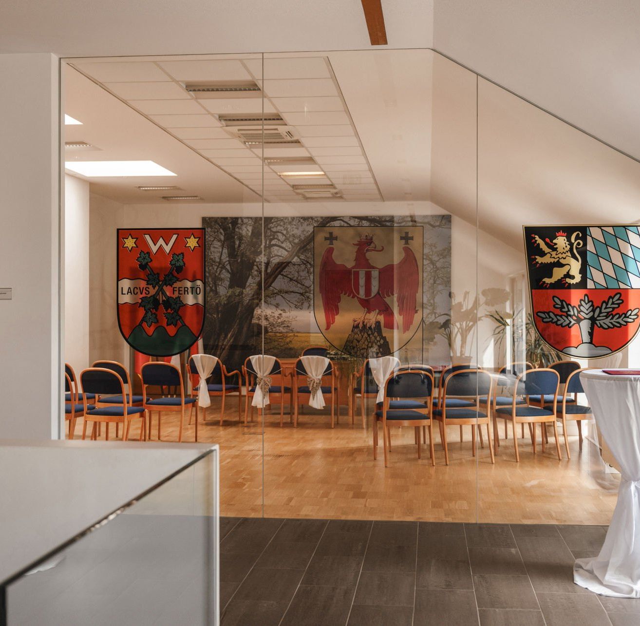 Ein moderner Bankettsaal mit blauen Stühlen und Holzboden. Flaggen hängen an der Wand und in der rechten Ecke befindet sich ein Tisch mit weißem Tuch.