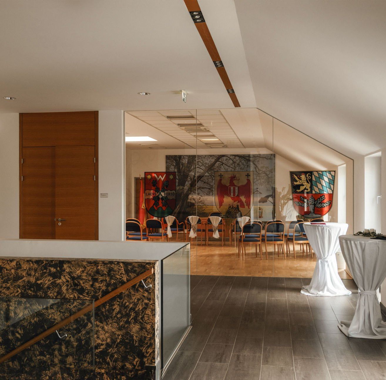 Ein großer Raum mit Holzböden und weißen Wänden. Ein Tisch mit einem weißen Tuch steht in der Ecke. Eine wandmontierte Lampe ist an der Wand angebracht. Einige Flaggen sind an der Wand montiert. Eine Tür befindet sich auf der linken Seite.
