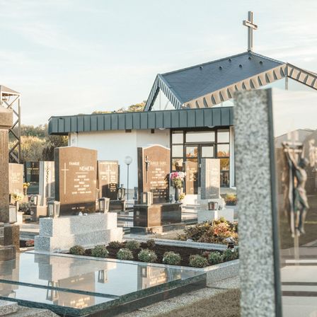 Ein Friedhof mit einer Kapelle, einem Kreuz und Grabsteinen. Die Familiennamen sind auf den Grabsteinen sichtbar, darunter Marie und Hemeth.