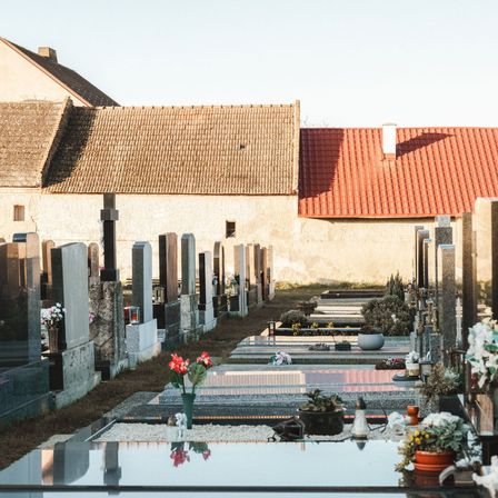 Ein Friedhof mit Grabsteinen und Gedenkstätten an einem sonnigen Tag. Ein Gebäude mit rotem Dach im Hintergrund.