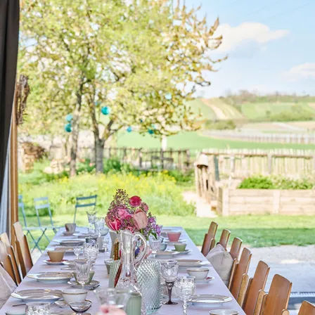 Ein langer Tisch mit Tellern, Gläsern und Blumen in einem Garten mit Blick auf ein grünes Feld und einen Baum.