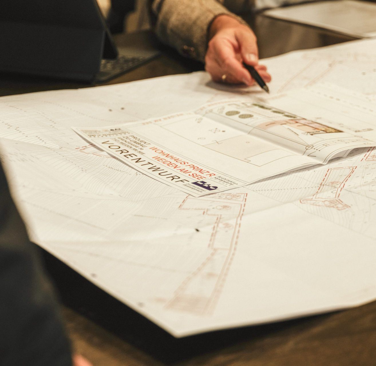 Eine Person zeigt auf einen großen architektonischen Bauplan mit roten Markierungen, eine Person im Hintergrund sitzt an einem Schreibtisch.