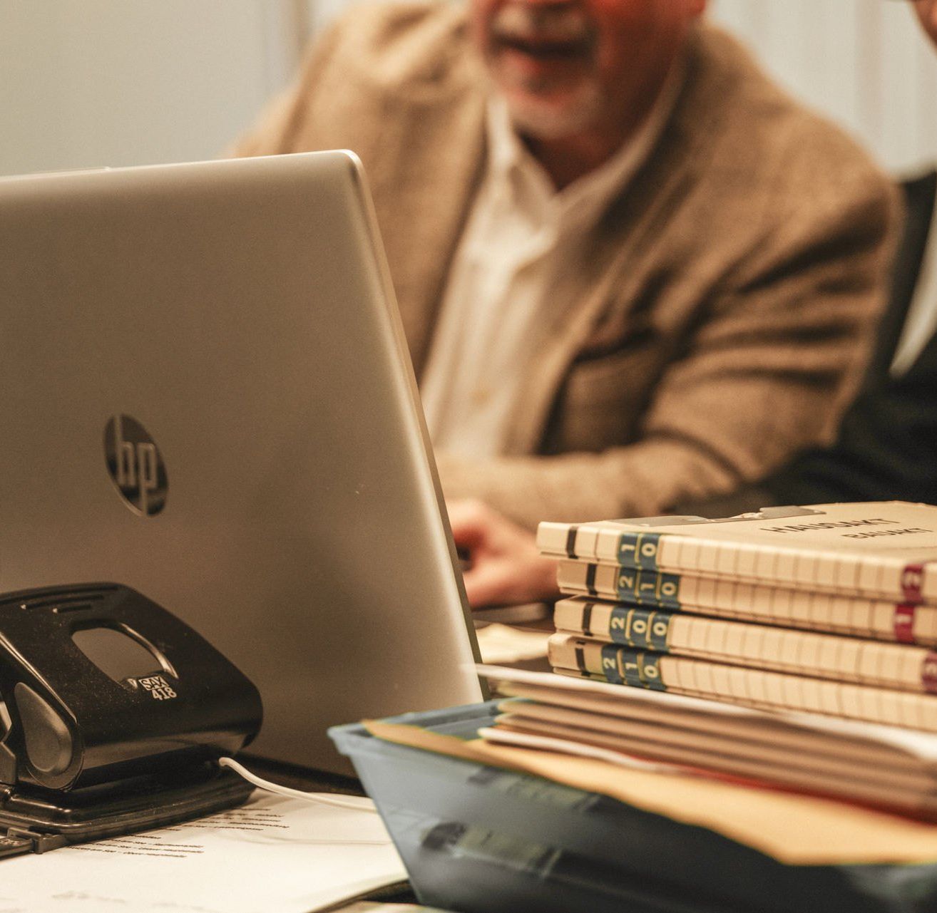 Zwei Personen, ein Mann und eine Frau, sitzen an einem Schreibtisch mit einem HP-Laptop und einem Stapel Notizbücher. Der Mann trägt einen beigen Mantel und die Frau einen schwarzen Mantel.