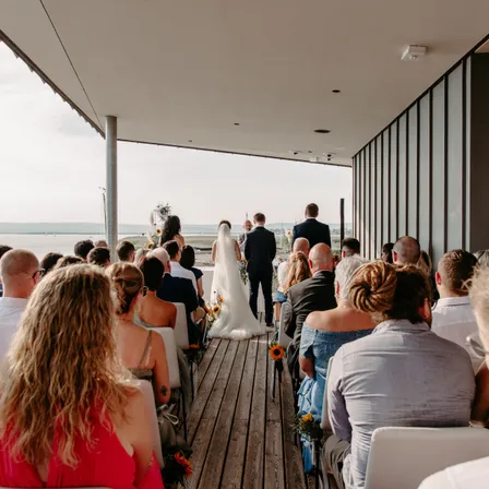 Eine Hochzeitszeremonie auf einer Terrasse mit Blick auf ein Gewässer. Die Braut in einem weißen Kleid und der Bräutigam in einem schwarzen Anzug gehen aufeinander zu. Gäste sitzen in Reihen und schauen den Paaren zu.