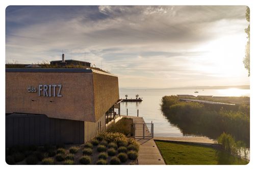 Ein Gebäude mit dem Wort Ritz befindet sich in der Nähe eines Gewässers mit einem Steg und Booten. Der Himmel ist bewölkt und es gibt einen Sonnenuntergang in der Ferne.