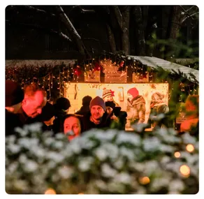 Eine Gruppe von Menschen steht bei Nacht vor einem Weihnachtsmarktstand, umgeben von Weihnachtslichtern und Bäumen. Einige tragen Mützen und Jacken.