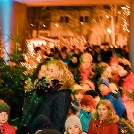 Eine Menschenmenge, einschließlich Kinder, steht bei Nacht vor Weihnachtsbäumen, mit festlichen Lichtern, die das Gebiet erhellen.
