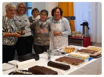 Eine Gruppe von Frauen steht hinter einem Tisch mit verschiedenen Desserts und lächelt für ein Foto. Der Tisch ist mit Schokolade, Sahne und anderen Gebäcksorten gefüllt.