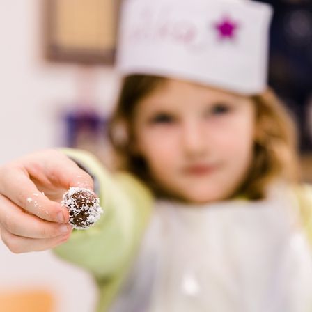 Ein junges Mädchen mit Kochmütze hält einen mit Kokosflocken überzogenen Schokoladentrüffel. Sie scheint für ein Foto zu posieren.