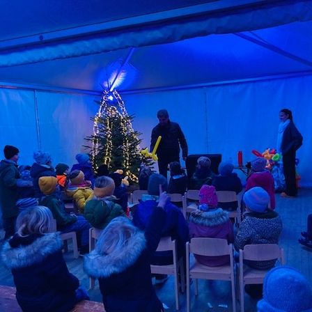 Ein Mann steht vor einem geschmückten Weihnachtsbaum und führt wahrscheinlich eine Darbietung für eine Gruppe von Kindern durch, die in Stühlen in einem blau beleuchteten Zelt sitzen.