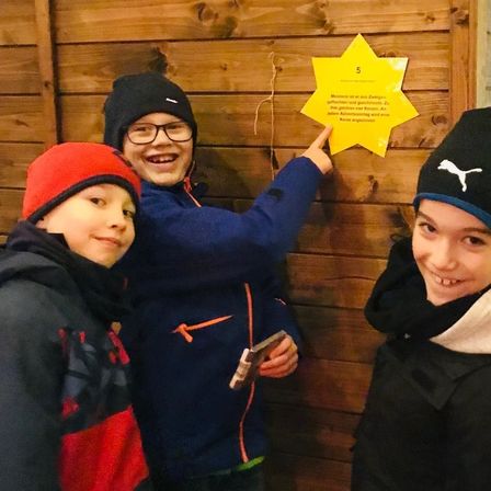 Drei Jungen stehen vor einer Holzwand mit einem gelben Stern, einer der Jungen zeigt darauf. Sie tragen Winterkleidung. Der Stern hat die Nummer 5 darauf.