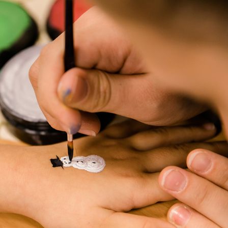 Eine Person malt mit einem Pinsel einen Schneemann auf die Hand einer anderen Person. Im Hintergrund sind verschiedene Farben sichtbar.