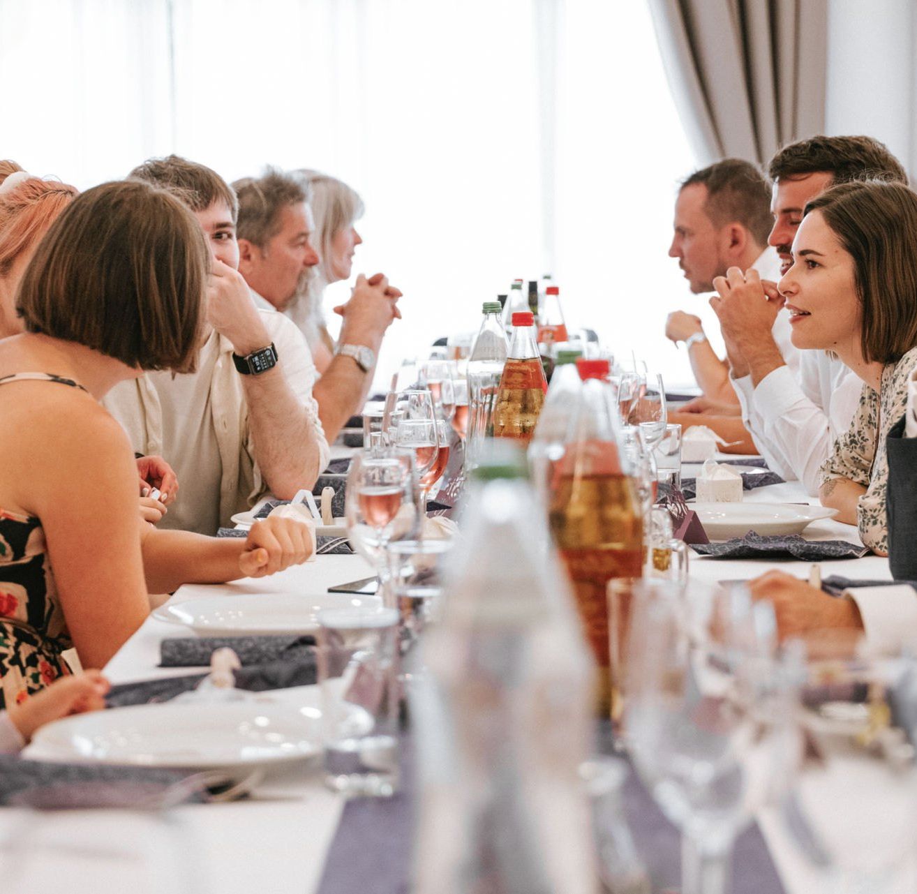 Eine Gruppe von Menschen sitzt in einem Restaurant an einem Tisch. Einige reden, andere essen und Weinflaschen stehen auf dem Tisch.