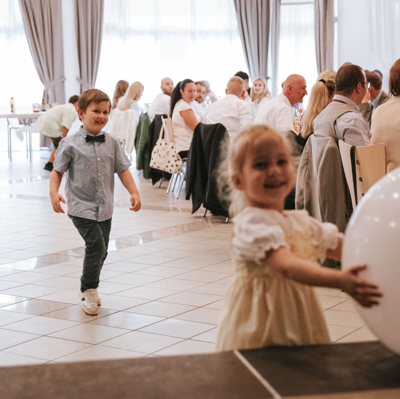 Ein Junge und ein Mädchen spielen mit großen weißen Luftballons in einem Indoor-Veranstaltungsraum mit Gästen, die an Tischen sitzen.