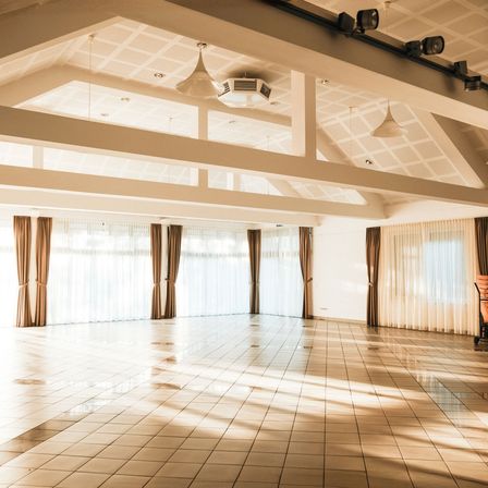 Ein leerer Saal mit großen Fenstern und Vorhängen, einem weißen Fliesenboden und einer Klimaanlage an der Decke.