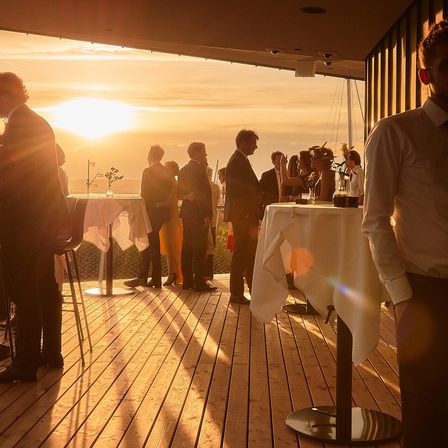 Auf einer Holzterrasse versammeln sich Menschen für eine Veranstaltung, während die Sonne untergeht und warmes Licht ausstrahlt. Einige stehen, andere sitzen, und Tische sind mit weißen Tüchern bedeckt. Auf der linken Seite ist eine Flagge zu sehen.