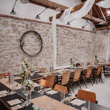 Ein rustikaler Essbereich mit Steinwand, Holzbalken und weißen Vorhängen. Eine runde Dekoration hängt an der Wand, und lange Tische sind mit Blumen, Kerzen und Weingläsern gedeckt.