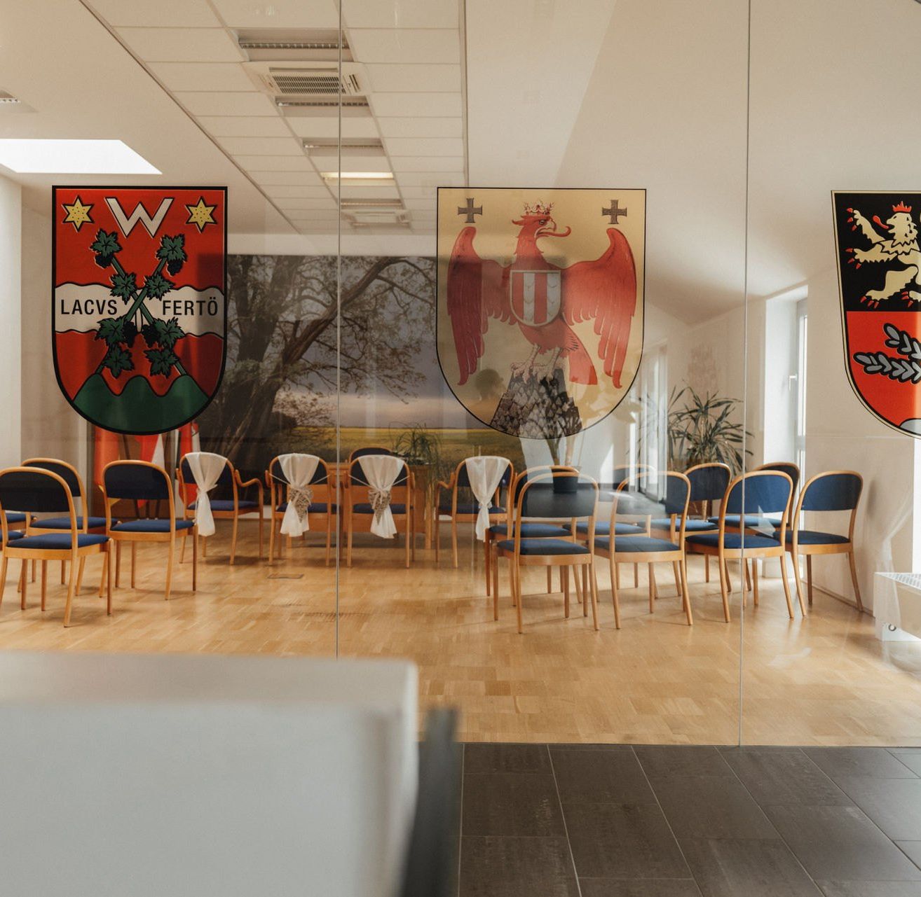 Ein Raum mit blauen Stühlen und weißen Tüchern darauf. Dahinter sind Glaswände mit Wappen und einem Baumbild. Der Boden ist aus Holz.