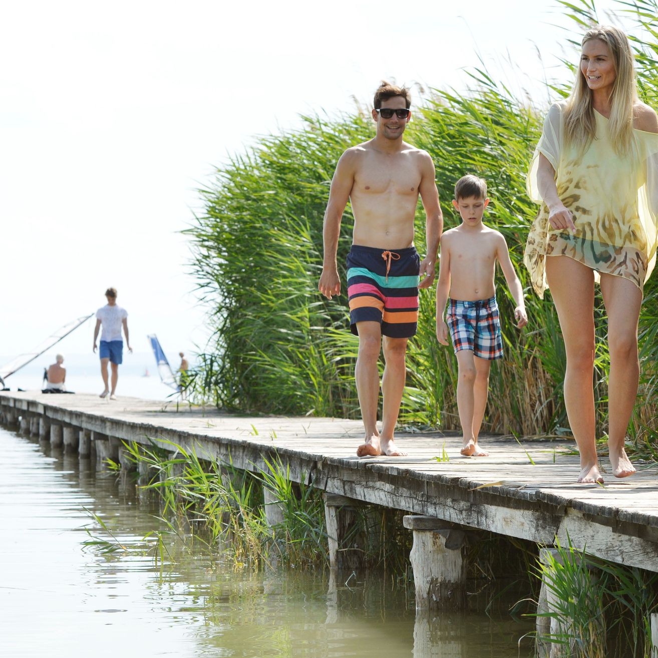 Eine vierköpfige Familie geht auf einem hölzernen Steg über das Wasser. Sie tragen alle Badekleidung.