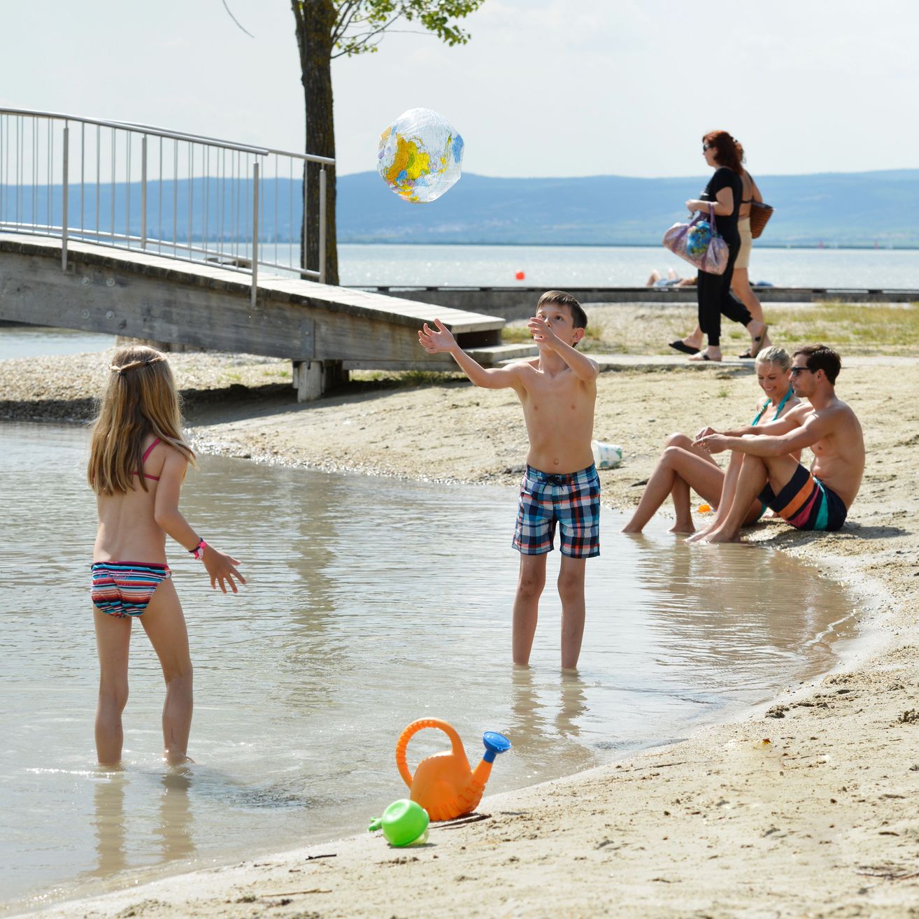 Ein Junge und ein Mädchen spielen am Strand, mit einem Paar im Hintergrund. Der Junge wirft einen Ball in die Luft, während das Mädchen zusieht. Links befindet sich eine Brücke und ein Baum.