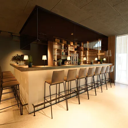 Eine moderne Bar mit schlankem Design, mit einem großen Tresen, Pflanzen, Lampen und Regalen mit Flaschen. Die Barhocker sind in einer Reihe angeordnet.