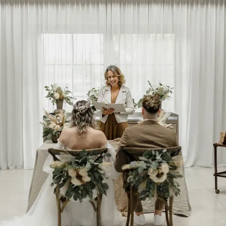 Eine Hochzeitszeremonie mit einer Braut und einem Bräutigam, die in Stühlen vor einem Tisch mit Blumen sitzen. Der Standesbeamte steht hinter dem Tisch, lächelt und liest aus einem Buch vor.