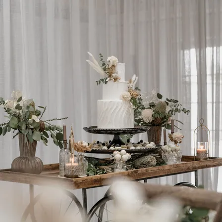 Ein weißer Hochzeitskuchen mit dekorativem Grün steht auf einem Holzwagen, umgeben von Blumenarrangements und Kerzen. Der Wagen steht vor einer weißen Gardine.