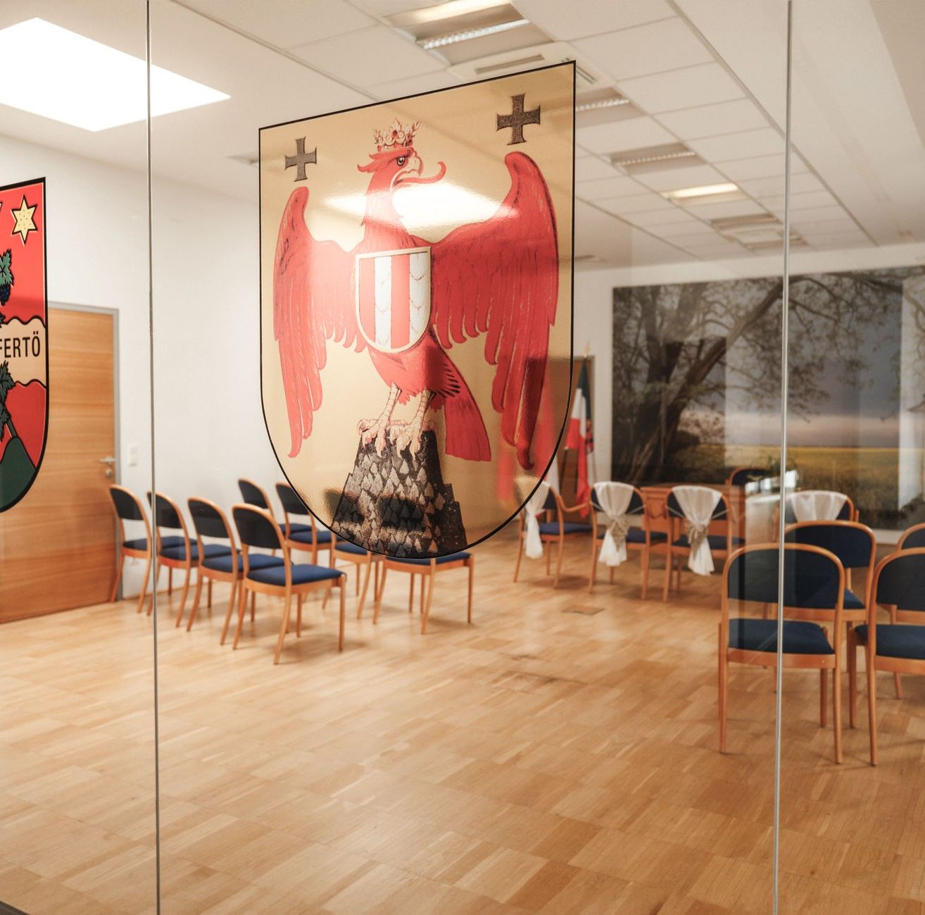 Ein Blick in einen Raum mit Holzboden und Glaswänden. An einer Wand befindet sich ein großes Wappen, vor dem Stühle aufgestellt sind. Ein weiteres Wappen befindet sich an der linken Wand.