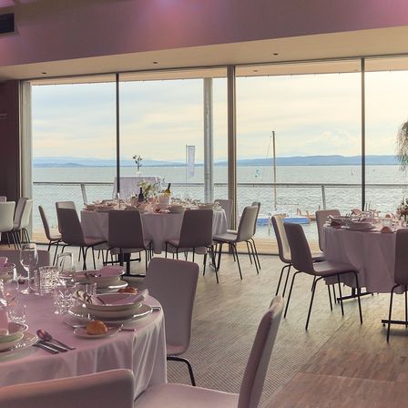 Ein Innenbereich für Mahlzeiten mit Tischen, die für eine Mahlzeit vorbereitet sind und Blick auf das Meer bieten. Die Tische sind mit weißen Tischdecken bedeckt und mit Gläsern, Tellern und Besteck angeordnet. Es gibt große Glasfenster, die den Ozean und die Berge zeigen.