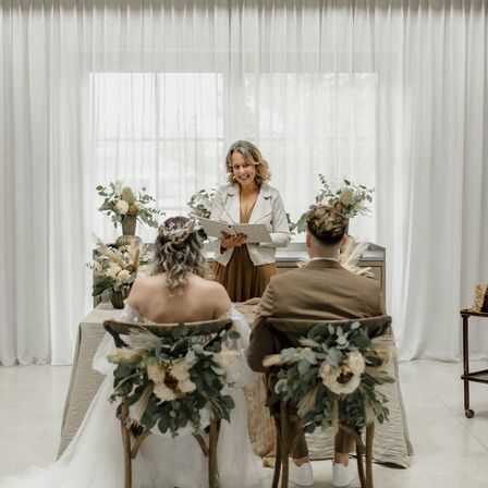 Eine Hochzeitszeremonie mit einer Braut und einem Bräutigam, die in Stühlen vor einem Tisch mit Blumen sitzen. Der Standesbeamte steht hinter dem Tisch, lächelt und liest aus einem Buch vor.