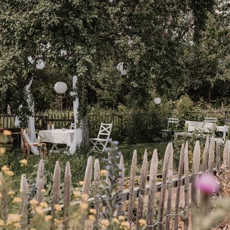 Ein Garten mit einem Tisch und Stühlen unter einem Baum, umgeben von hängenden Laternen. Der Garten ist von einem Holzzaun umgeben und hat viele Pflanzen und Blumen.