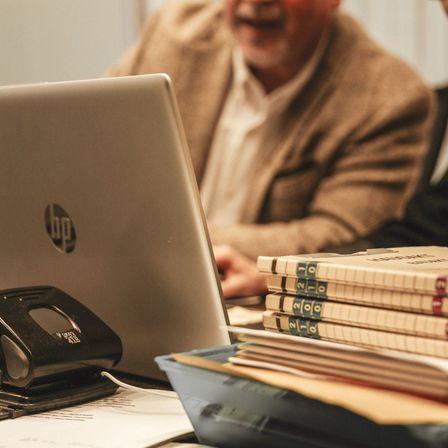 Ein Mann sitzt an einem Schreibtisch mit einem Laptop und einem Stapel Notizbüchern. Der Laptop ist geöffnet, und die Notizbücher sind ordentlich gestapelt.