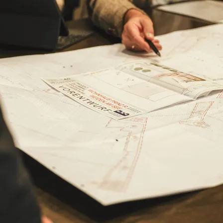 Eine Person arbeitet an architektonischen Plänen auf einem großen Tisch, mit einem Buch, das auf einer Seite mit dem Titel 'Lorentzwurp' geöffnet ist.