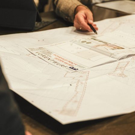 Eine Person arbeitet an architektonischen Plänen auf einem großen Tisch, mit einem Buch, das auf einer Seite mit dem Titel 'Lorentzwurp' geöffnet ist.