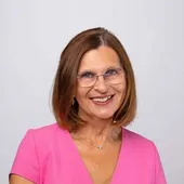 Eine Frau mit einem rosa Oberteil und einer Brille lächelt und posiert für ein Foto. Sie hat kurze Haare und eine Halskette mit einem Anhänger. Der Hintergrund ist weiß.