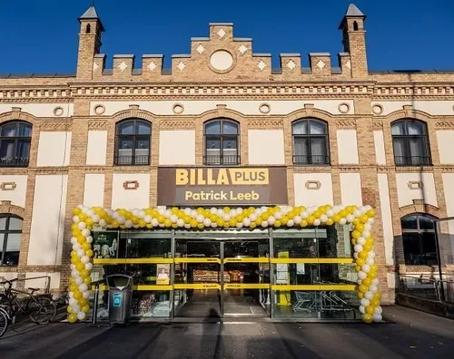 Der Vordereingang eines BILLA PLUS-Geschäfts ist mit gelben und weißen Luftballons geschmückt. Das Gebäude hat eine Backsteinfassade mit Bogenfenstern. Links vom Eingang sind Fahrräder geparkt.