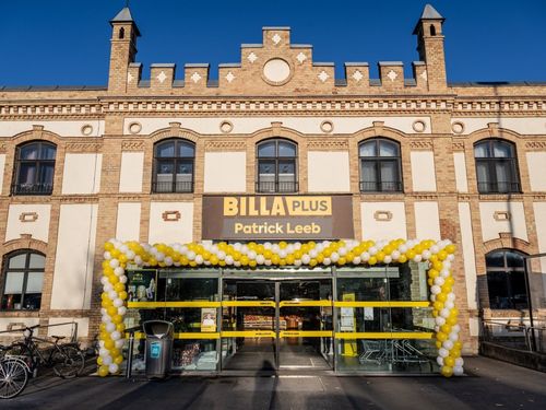 Ein großes Gebäude mit einem Schild, das BILLA PLUS anzeigt. Vor dem Eingang befindet sich ein gelber und weißer Luftballonbogen. Der Laden hat eine Glastür und Fenster. Auf der linken Seite des Eingangs steht ein Mülleimer.
