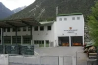 Ein weißes Gebäude mit einem grünen Dach und mehreren Fenstern steht vor einem Berg, mit einer großen offenen Garagentür und einem Metallzaun davor.